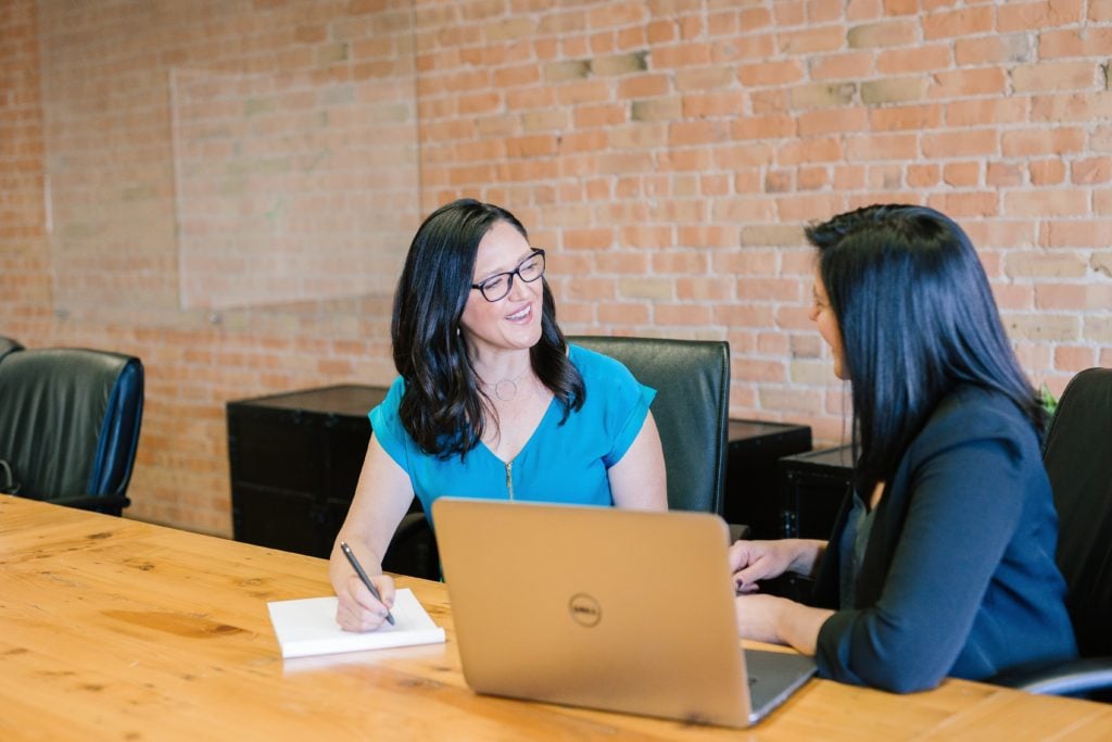 two women talking on a table with a computer taking notes