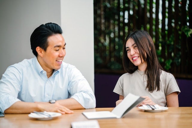 two people seated side by side and smiling. there are papers on a wooden table in front of them